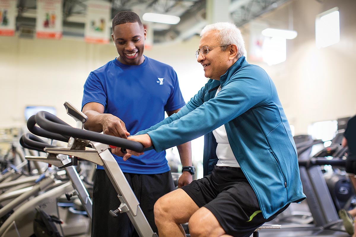 A fitness trainer assists an older man using a stationary bike in a gym, both smiling during the session.