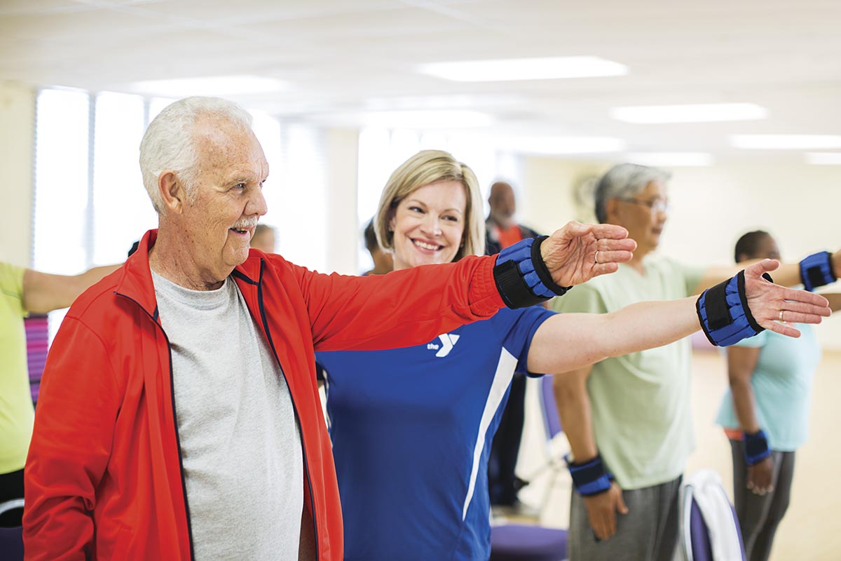 Older adults exercising in a group fitness class with wrist weights, guided by a smiling YMCA instructor in a bright, spacious room.