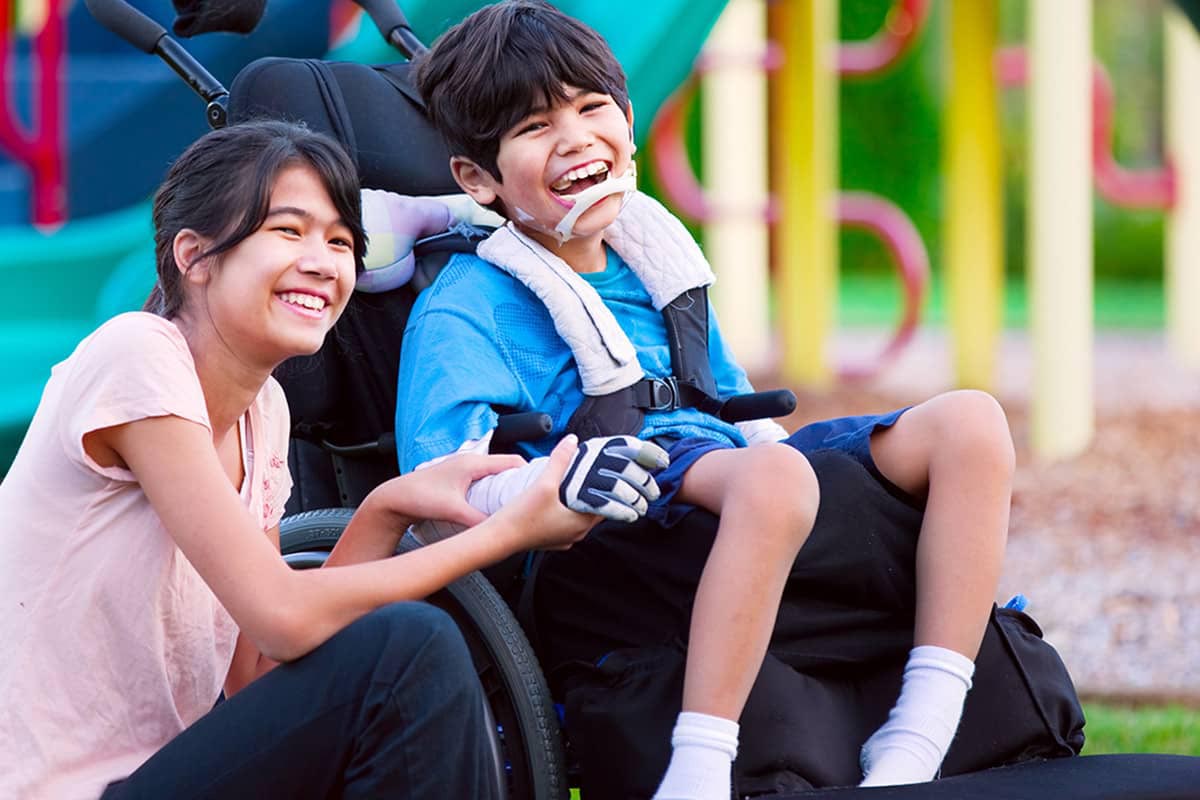 Smiling boy in a wheelchair and a young girl laughing together at a playground, surrounded by colorful equipment.