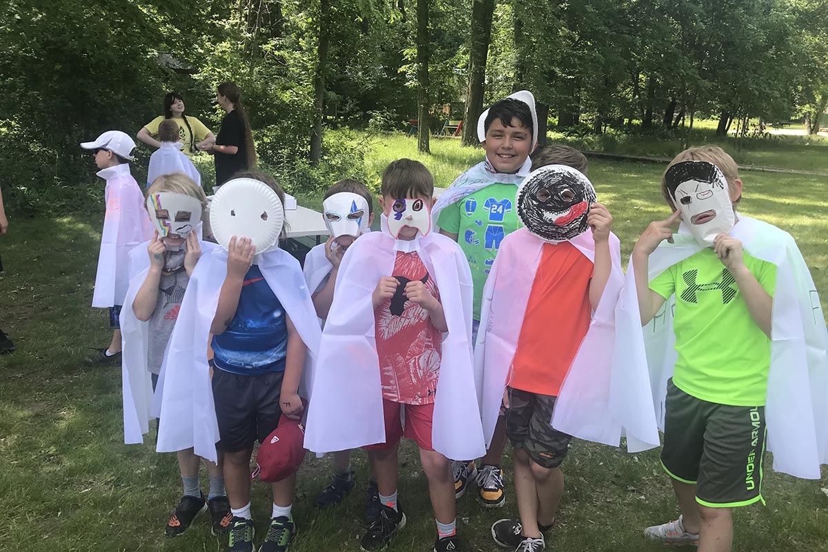 Children outdoors wearing handmade paper masks and white capes, participating in a creative activity during a summer camp.