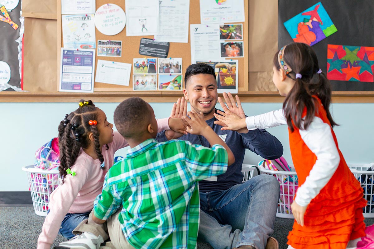 A smiling teacher sitting on the floor giving high-fives to a group of children in a colorful classroom with bulletin board artwork in the background.