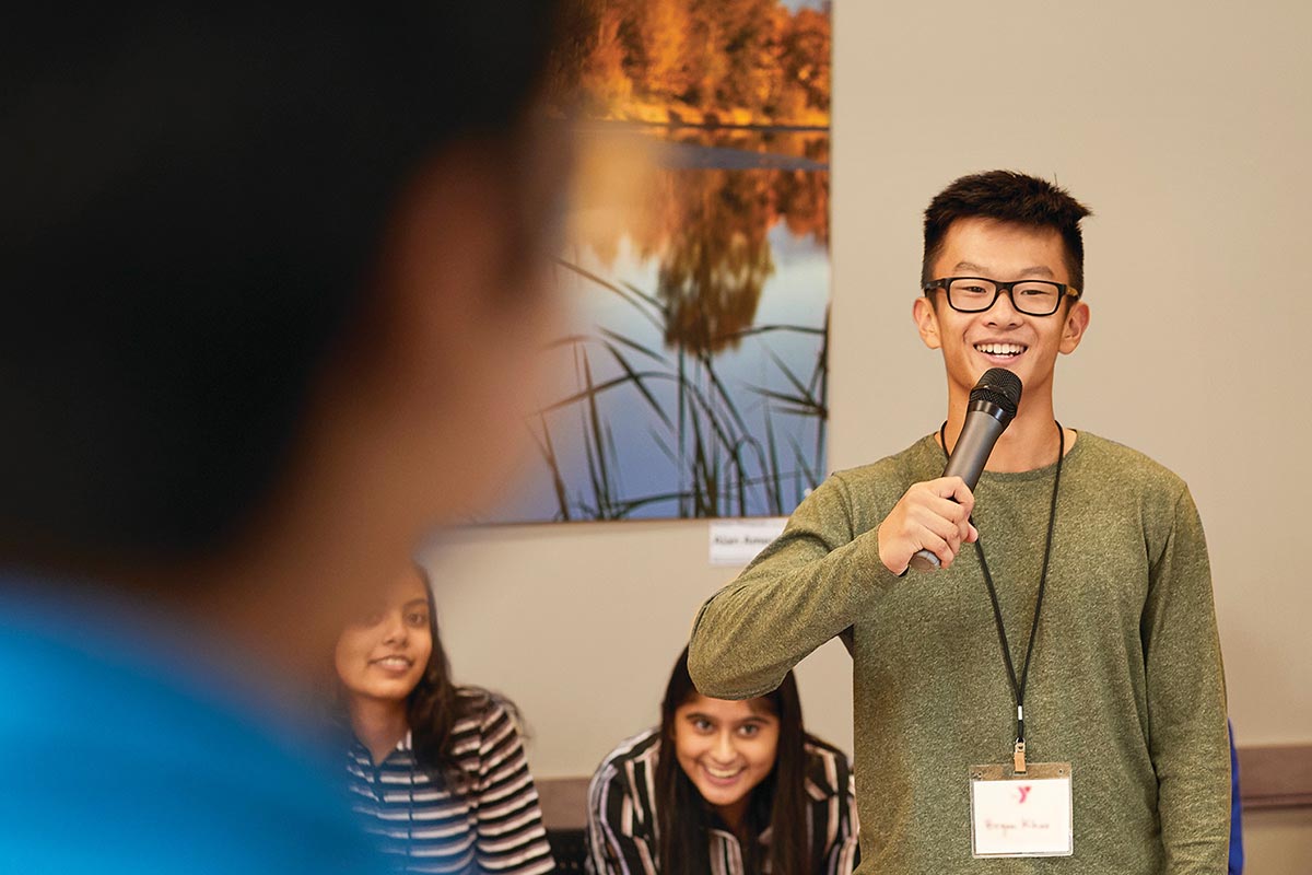 A young man wearing glasses smiles while speaking into a microphone during a group activity, with others seated and listening in the background.