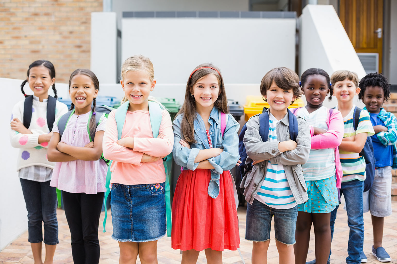 Group of young children standing outdoors with backpacks, smiling confidently with arms crossed, in front of a school building.