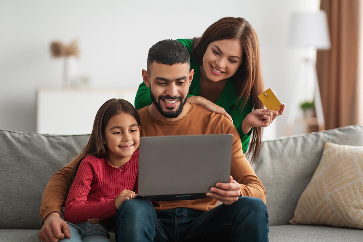 Smiling family sitting on a couch, using a laptop together; the mother holds a credit card, suggesting online shopping or learning.