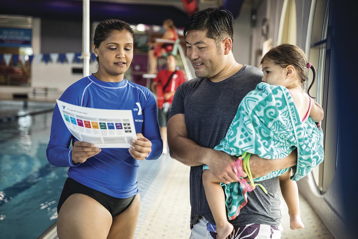 Swim instructor in a blue uniform discussing a flyer with a parent holding a young child wrapped in a towel near an indoor pool.