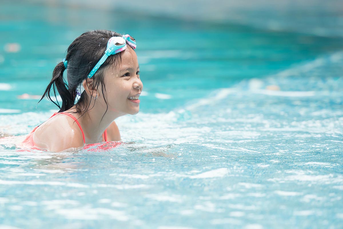 Young girl wearing swim goggles smiling while swimming in a pool, enjoying water activities.