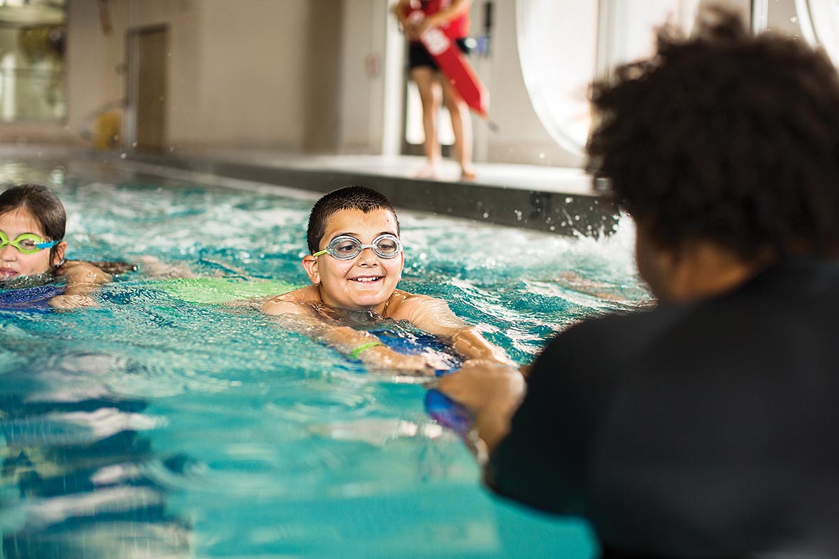 Child in goggles smiling during a swim lesson in an indoor pool, instructor guiding from the water.