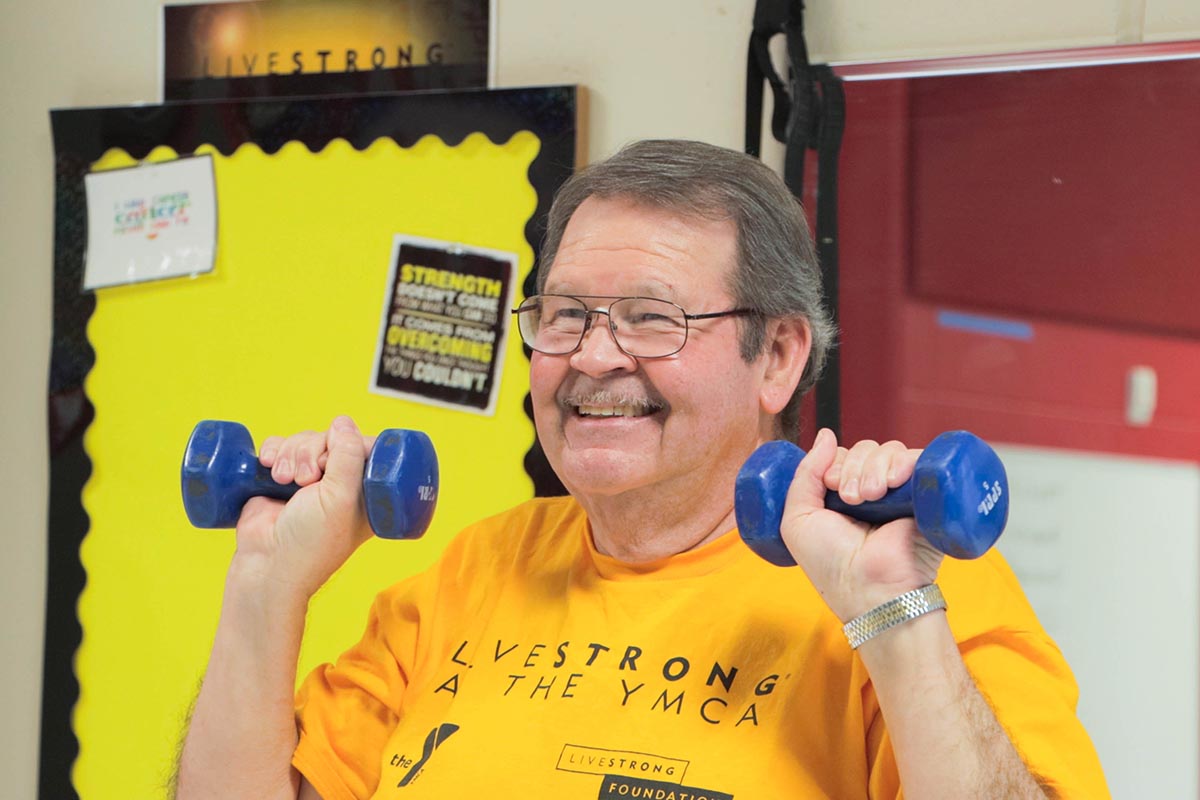 Older man smiling while lifting blue dumbbells in a fitness class, wearing a yellow Livestrong at the YMCA t-shirt, with a motivational bulletin board in the background.