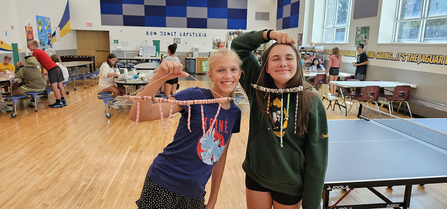 Two girls proudly displaying their handmade bead crafts in a school cafeteria, surrounded by other kids engaged in activities at tables.