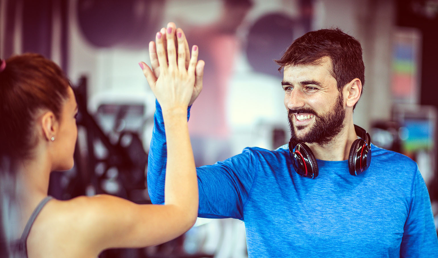 A man and woman giving a high-five in a gym, smiling and celebrating teamwork or success. The man wears a blue shirt and headphones around his neck, with blurred gym equipment in the background.