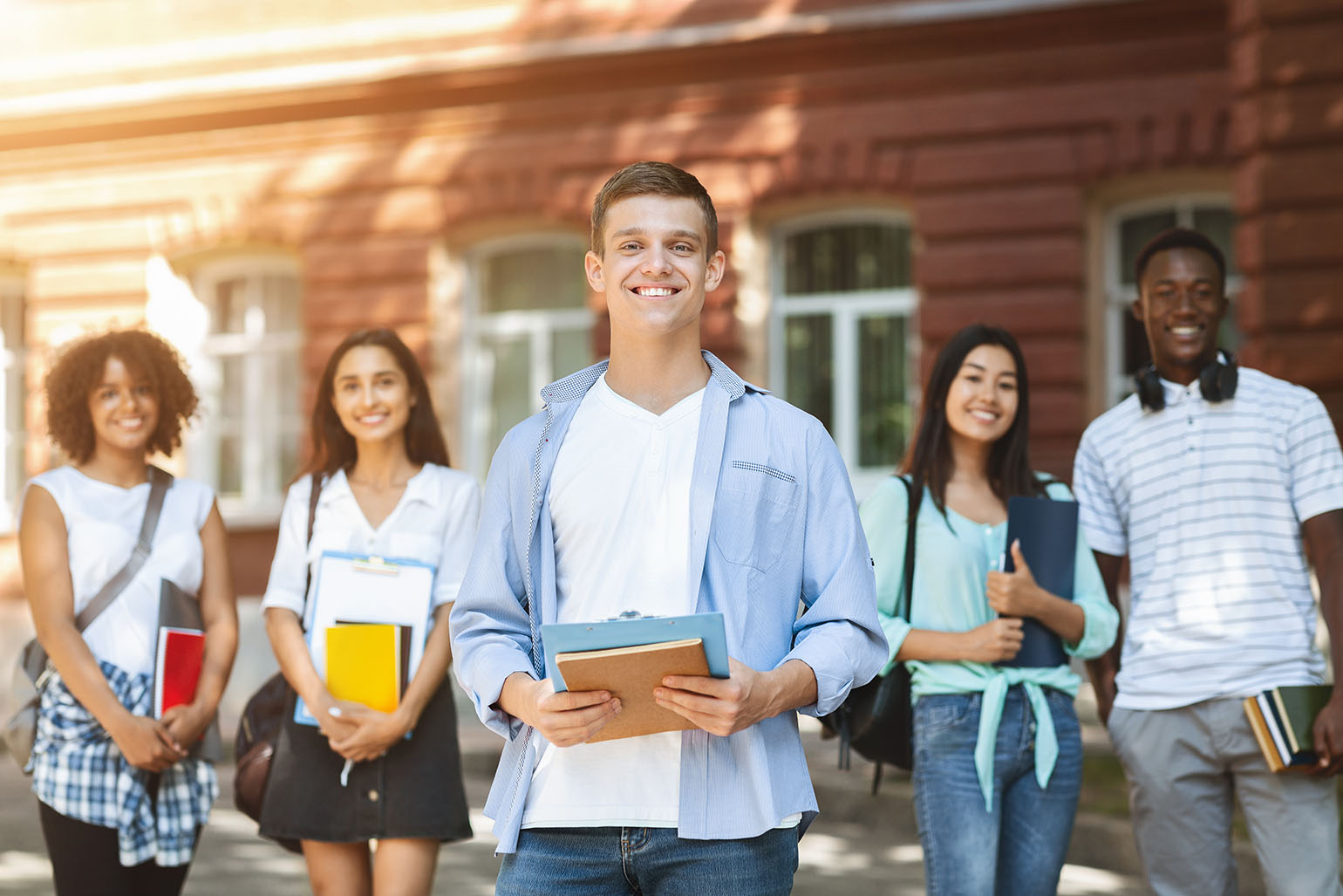 Group of diverse students smiling outdoors, holding books and notebooks, standing in front of a school building.