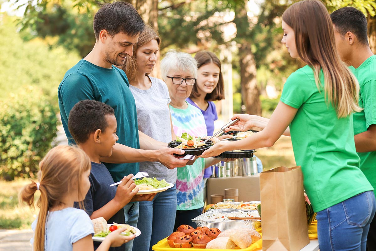 Volunteers serving meals to a diverse group of people outdoors, with a table of food trays, bread, and muffins in the foreground.