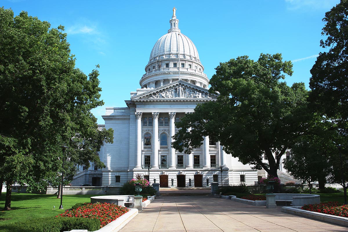 Front view of a state capitol building with a domed roof, surrounded by trees and landscaped gardens under a clear blue sky.