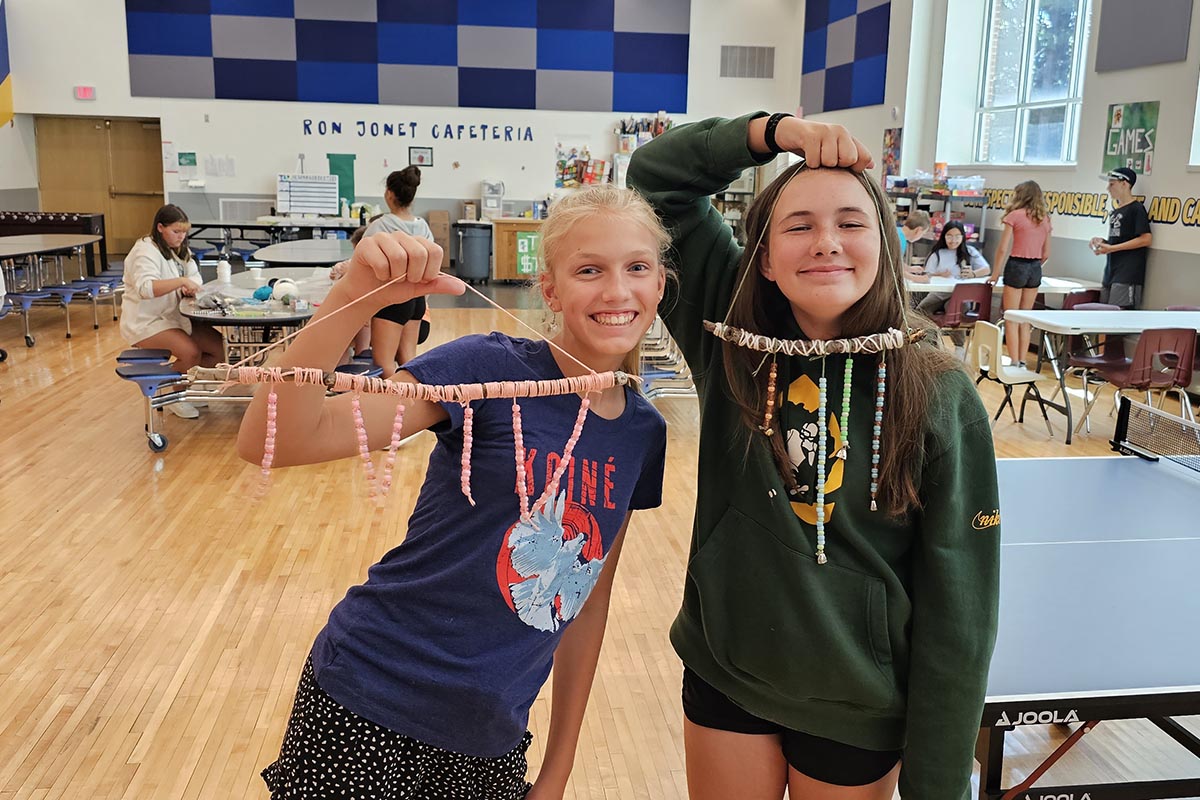 Two girls in a cafeteria proudly holding handmade bead and string crafts, smiling at the camera.