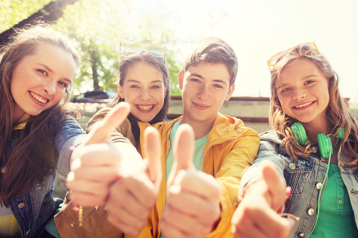 Four smiling teenagers giving thumbs-up outdoors on a sunny day.