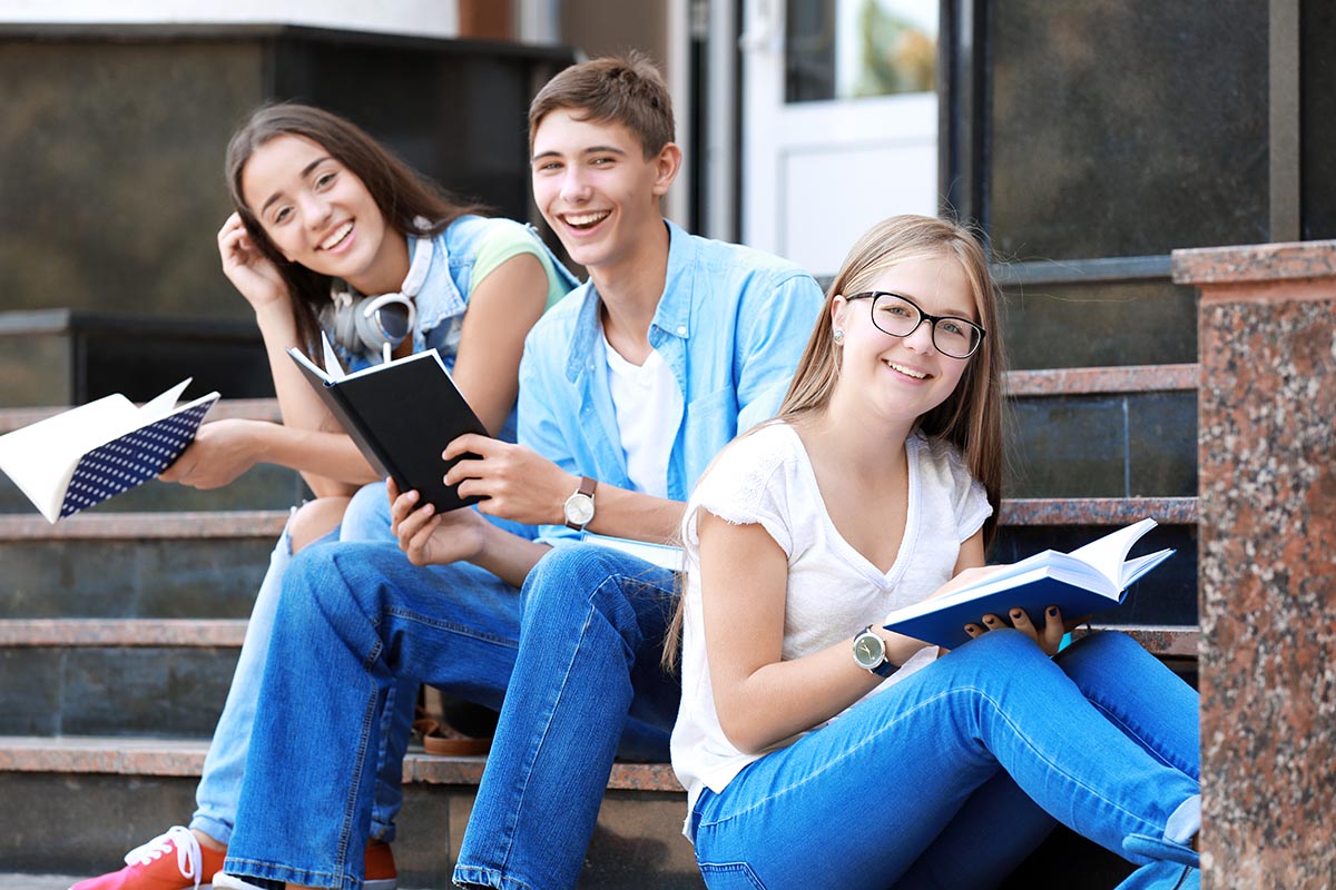 Three smiling students sitting on outdoor steps, holding books, and studying together.