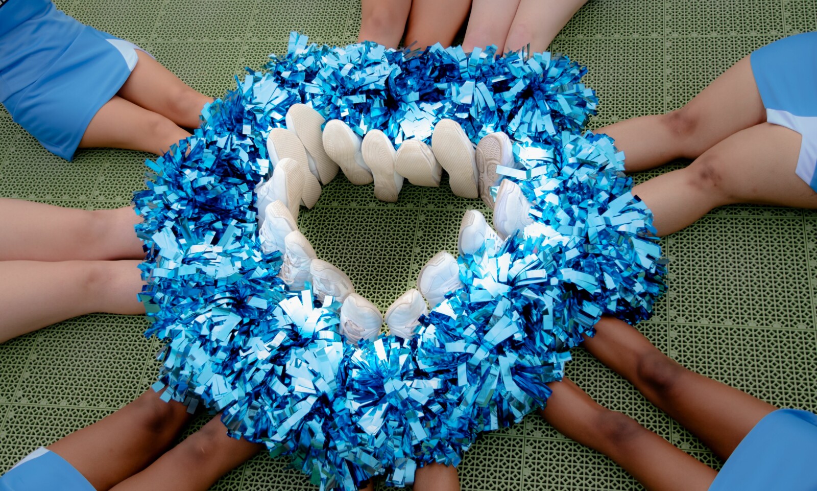 Cheerleaders sitting in a circle, their white sneakers and hands holding blue pom-poms forming a vibrant centerpiece on a green mat.